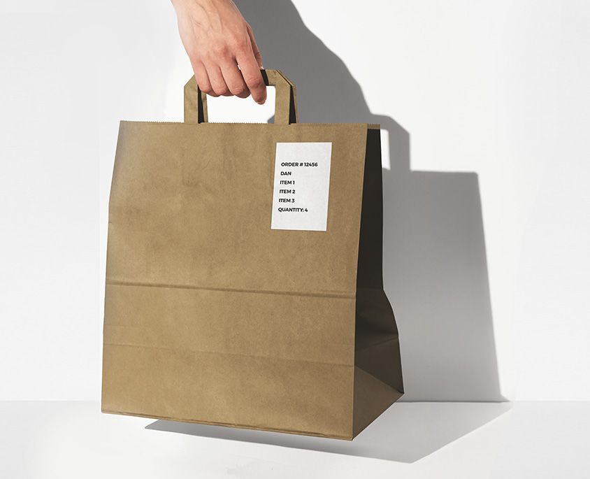 Hand holding a brown paper takeout bag with a printed order label showing order number, name, and items